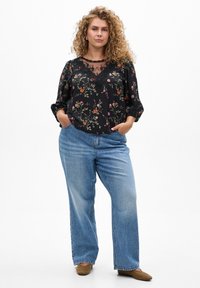 Žena s kovrčavom plavom kosom koja nosi crnu cvjetnu čipkastu bluzu, plave široke traperice i smeđe cipele, stoji s rukama u džepovima.