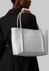 Borsa tote bianca in pelle con due manici lunghi, dotata di tasca frontale e linee pulite. Il design è minimalista con una texture liscia.