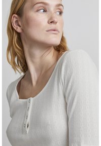Hvid strikket top med et tekstureret mønster, rund halsudskæring og placket med tre knapper. Ærmer til albuen og tætsiddende design.
