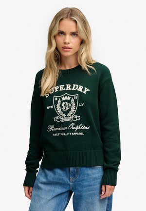 Mujer rubia con un suéter Superdry verde oscuro con texto blanco y vaqueros azules, posando frente a un fondo claro y liso.