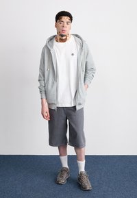 Sudadera gris con cordones, camiseta blanca, pantalones cortos grises y zapatillas grises. Tejido texturizado, diseño casual y logo visible en la camiseta.