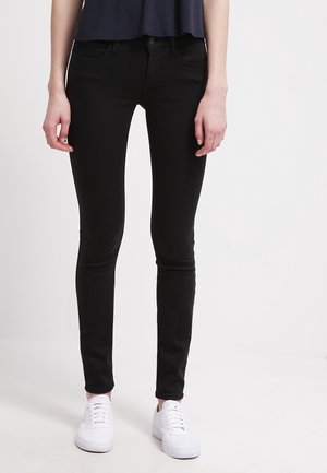 Personne portant un jean skinny noir, un haut bleu marine et des baskets blanches, debout sur un fond blanc.