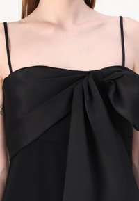 Robe noire à fines bretelles, ornée d'un grand nœud au niveau de la poitrine et d'une texture lisse et fluide. Design simple et élégant.