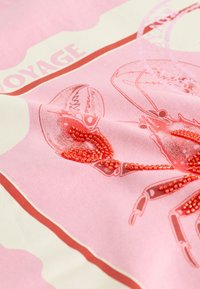 Lyserødt tekstil med krabbeprint og røde perlepynt. Designet inkluderer tekst, med kontrasterende cremefarvede sektioner og subtile mønstre.