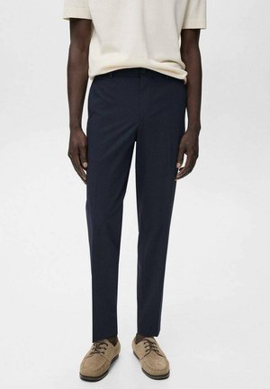 Pantaloni su misura blu navy con taglio dritto, realizzati in un tessuto morbido, abbinati a scarpe stringate color marrone chiaro.