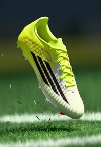 Κίτρινο ποδοσφαιρικό παπούτσι με υφή και μαύρη ρίγα, διαθέτει κορδόνια και κόκκινη λεπτομέρεια, που αιωρείται πάνω από γρασίδι.