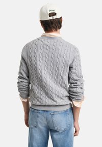 Grå cable-knit sweater med rund halsudskæring, ribstrikkede manchetter og kant. Blød tekstur. Båret over en lys, stribet skjorte og parret med blå jeans.