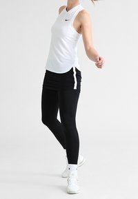 Biała bezrękawnik sportowy z wysokim kołnierzem oraz czarnym logo Nike, w połączeniu z czarnymi legginsami z białymi akcentami po bokach oraz białymi sneakersami.