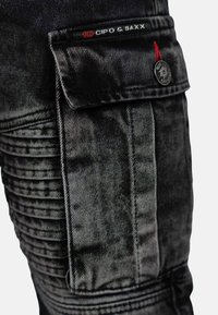 Black denim cargo pants with textured ribbing on sides, a flap pocket, and a logo tag. Features silver hardware and a fitted design.