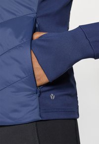 Tamsiai mėlynas sportinis marškinėlis su užtrauktuko kišenėle. Turi lygią tekstūrą, prigludusį dizainą ir logotipo detalę apačioje.