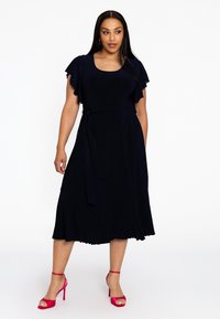 Navyblaues Kleid mit flatternden Ärmeln, einem Taillengürtel und einem ausgestellten Saum. Kombiniert mit knallpinken High-Heels. Glatte Stofftextur.