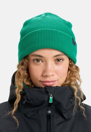 Groene gebreide beanie met ribbelstructuur, voorzien van een klein logo detail. Ggedragen met een zwarte ritsjack met hoge kraag.
