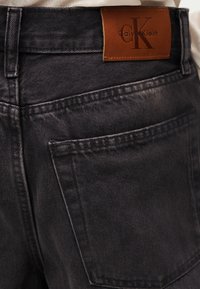 Πίσω τσέπη μαύρου τζιν με καφέ δερμάτινο πλακέ με το λογότυπο Calvin Klein ραμμένο πάνω από την τσέπη.