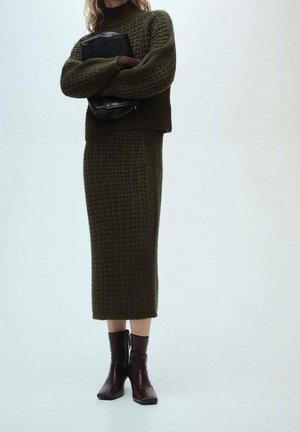 Femme portant un pull en maille vert olive et une jupe midi assortie, tenant une pochette en cuir noir, associée à des bottines marron foncé.