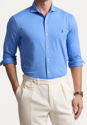 Man draagt een nauwsluitend hemelsblauw overhemd met opgerolde mouwen, ingestopt in hoogtaill broek met crèmekleurige plooien, en een bruine leren horloge.