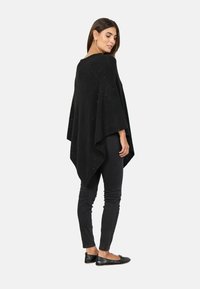 Sort poncho lavet af tekstureret stof, med en løs pasform og asymmetrisk kant. Sammen med sorte skinny jeans og flade sko.