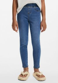 Úzké džíny ve středně modrém denimu, s klasickým designem pěti kapes, prošitím a hladkou texturou, doplněné béžovými sandály.