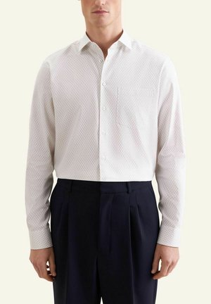 Mann trägt ein weißes Hemd mit dezentem Muster und Brusttasche, kombiniert mit schwarzen, plissierten Hosen.