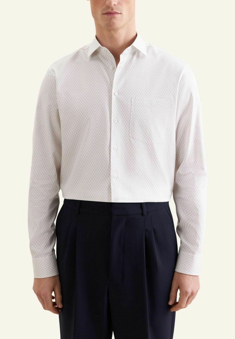 Homme portant une chemise blanche boutonnée avec un motif subtil et une poche poitrine, associée à un pantalon noir plissé.