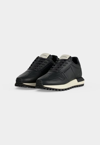 Paire de baskets basses noires avec semelles blanches et lacets noirs sur un fond clair uni.