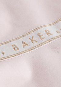 Rosa Stoff mit einem weißen Etikett, auf dem "BAKER" in goldenem Faden gestickt ist. Die Textur ist glatt und weich.