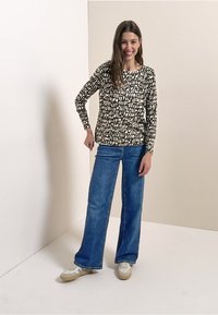 Schwarz-weiß gemustertes Langarmshirt kombiniert mit blauen, weit geschnittenen Jeans. Lässige Schuhe vervollständigen das Outfit. Heller Hintergrund.