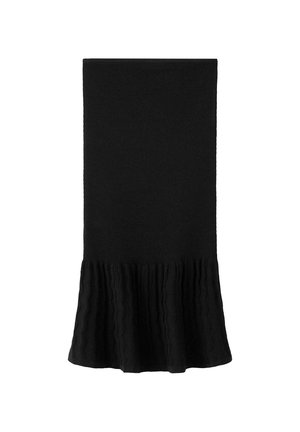 Giovane donna in piedi che indossa un abito nero senza spalline con orlo strutturato, sandali marroni borchiati e porta una piccola borsa marrone a tracolla.