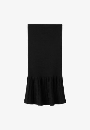 Giovane donna in piedi che indossa un abito nero senza spalline con orlo strutturato, sandali marroni borchiati e porta una piccola borsa marrone a tracolla.