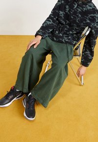 Schwarze Tarnfleecjacke, lockere olivgrüne Hose, schwarze und rote Sportschuhe. Sitzend auf einem Metallstuhl vor einem gelben Bodenhintergrund.