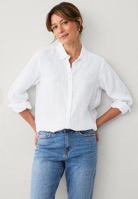 Camicia button-up bianca in lino con colletto, maniche arrotolate e tessuto leggero, abbinata a jeans in denim blu.