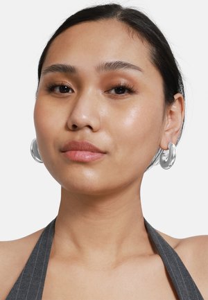 Orecchini a cerchio in argento con un design liscio e curvo. Il modello ha i capelli neri e lisci e indossa un top a halter grigio a righe.