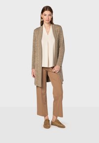 Stickad kappa, beige och texturerad, öppen fram, kombinerad med en krämfärgad topp och bruna vida byxor; bruna snörskor fulländar looken.