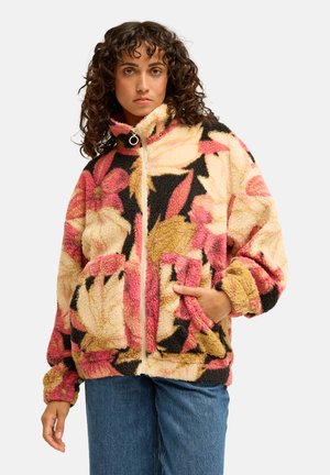Woman with curly dark hair wearing a colorful floral patterned zip-up fleece jacket and blue jeans, one hand in pocket, neutral expression.