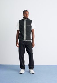 Mand der står på blå gulv iført sort vest over hvid t-shirt, sorte bukser og hvide sneakers, som ser lidt til siden.