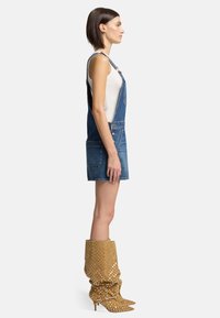 Abito in denim blu con bretelle regolabili, dotato di tasche frontali. Abbinato a stivali gialli a pois, alti fino al ginocchio, con punta affusolata.