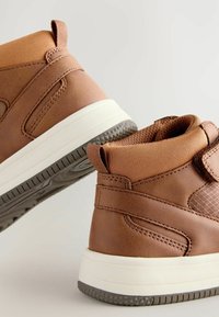 Brązowe skórzane trampki typu high-top z teksturowanym brązowym panelem, elastycznymi akcentami, białą podeszwą i gumową przyczepnością na zewnętrznej podeszwie.