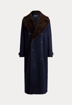 Femme aux longues tresses portant un manteau bleu marine à double boutonnage avec un grand col en fourrure, une ceinture décorative marron, un pantalon gris et des chaussures marron, debout immobile.