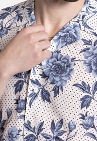 Man in een beige shirt met een blauw bloempatroon en zwarte stippen, hand die de kraag aanraakt, toont een deel van een gouden ketting en baard.