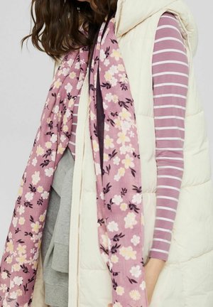 Personne portant un gilet matelassé crème, une chemise à manches longues rayée rose et blanche, et une écharpe fleurie rose sur un haut gris, montrée des épaules à la taille.