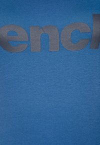 Modrá bavlněná tričko s velkým černým písmem, které hlásí "French." Textura je hladká s mírným strečem.