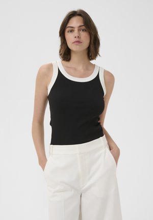 Kobieta z brązowymi włosami do ramion, ubrana w czarny top bez rękawów z białym wykończeniem i białe spodnie z wysokim stanem, ręce w kieszeniach.