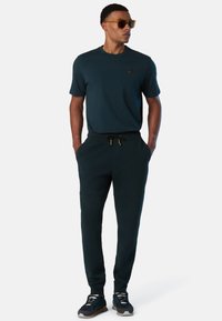 Hombre vestido con un conjunto casual de color azul oscuro, con camisa de manga corta, pantalones jogger con cordón, zapatillas y gafas de sol, de pie con las manos en los bolsillos.