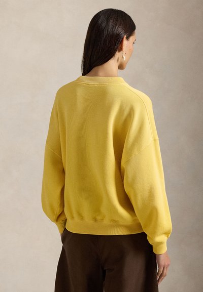 Κίτρινο φούτερ με άνετη εφαρμογή, ριμπ μανσέτες και τελείωμα, και χαμηλωμένους ώμους. Ορατή είναι η απαλή υφή και οι ελάχιστες ραφές.