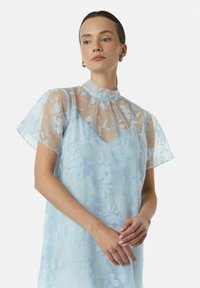 Jasnoniebieska sukienka z przezroczystą kwiatową nakładką, wysokim kołnierzem, krótkimi falbanistymi rękawami oraz luźnym krojem. Delikatne wzory kwiatowe na całej powierzchni.
