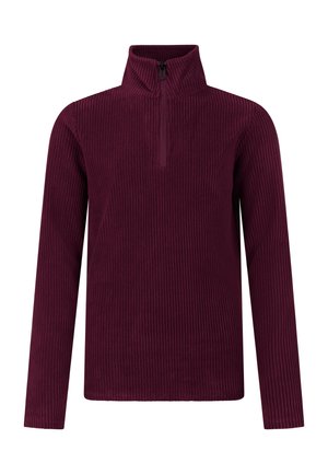 Jonge vrouw die een magenta lange-mouwen top draagt, een roze-beige muts, een broek met luipaardprint en zwarte schoenen, staand tegen een effen achtergrond.