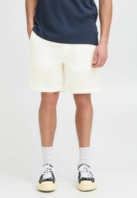 Light beige woven shorts with a textured pattern, featuring side pockets and a straight cut, paired with black and cream sneakers and white socks.