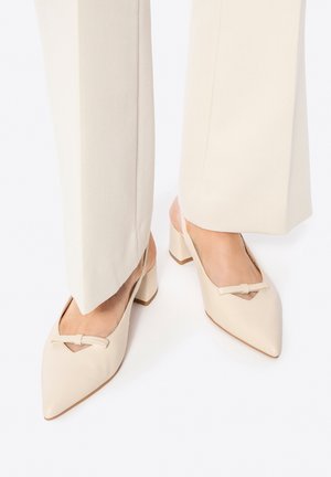Pies con zapatos de tacón crema de punta puntiaguda con tiras traseras, tacones anchos y un pequeño detalle de lazo, combinados con pantalones de pierna ancha en beige claro.