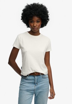 Mujer con cabello rizado que lleva una camiseta blanca de manga corta metida en unos vaqueros azul claro con un cinturón marrón trenzado, de pie sobre un fondo blanco.