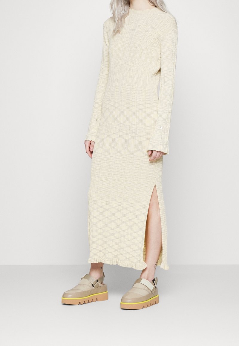 Femme portant une robe en maille crème à manches longues avec une fente latérale et des chaussures plateformes beige à semelles jaunes, debout devant un fond uni.