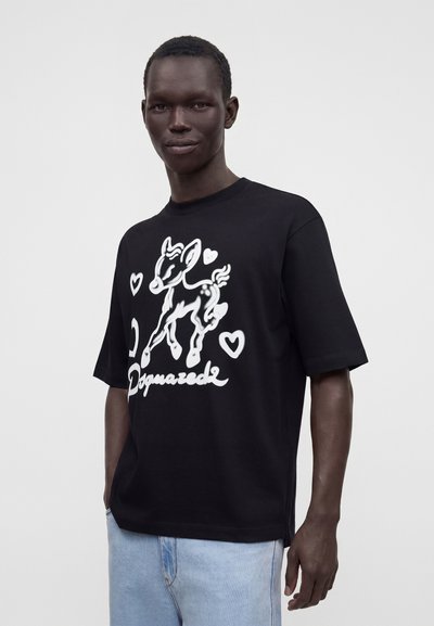 Άνδρας που φοράει μαύρο φαρδύ t-shirt με λευκό σχέδιο ελαφιού και καρδιών και ανοιχτό μπλε τζιν μπροστά σε απλό λευκό φόντο.